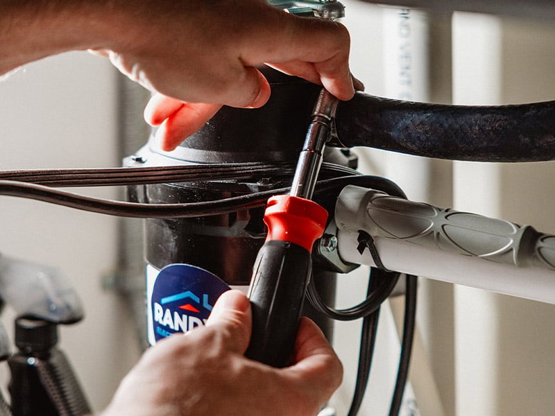 Hands using a screwdriver to adjust pipes under a kitchen sink.